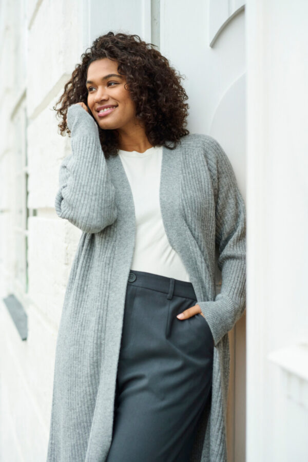 Frau lehnt an der Wand und trägt einen langen grauen Cardigan über einem weissen Tshirt und einer grauen Hose