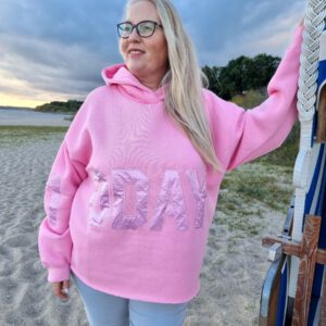 Blonde Frau steht an Strandkorb gelehnt am Strand und trägt einen pinken oversized Hoodie mit Aufdruck auf rmeln und Bauch von 10days