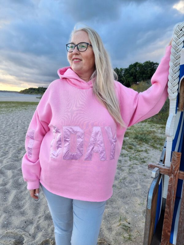 Blonde Frau steht an Strandkorb gelehnt am Strand und trägt einen pinken oversized Hoodie mit Aufdruck auf rmeln und Bauch von 10days