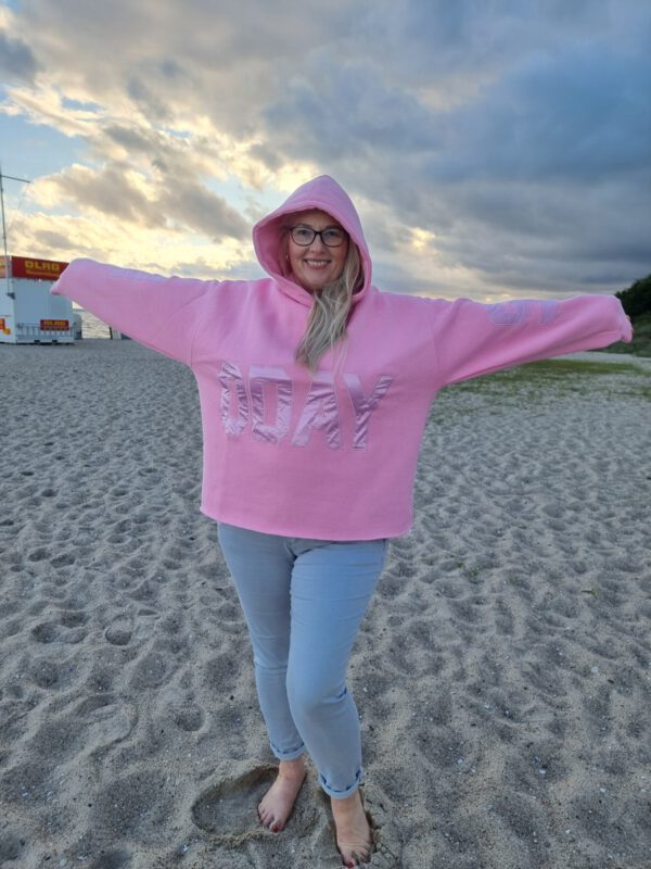 Frau in Ganzfigur steht am Strand und trägt einen Hoodie mit Kapuze und Aufdruck an Ärmeln und im Bauchbereich 10days