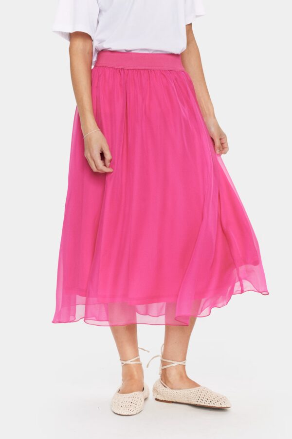 Langer, pinkfarbener Damenrock aus leichtem Chiffon, mit elastischem Bund, getragen mit weißem T-Shirt und flachen Espadrilles, vor weißem Hintergrund